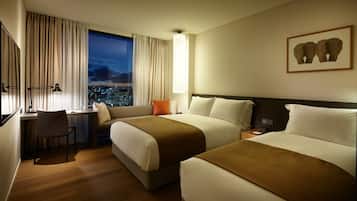 Habitación de hotel con dos camas, un escritorio, una silla y un amplio ventanal con vista a la ciudad.
