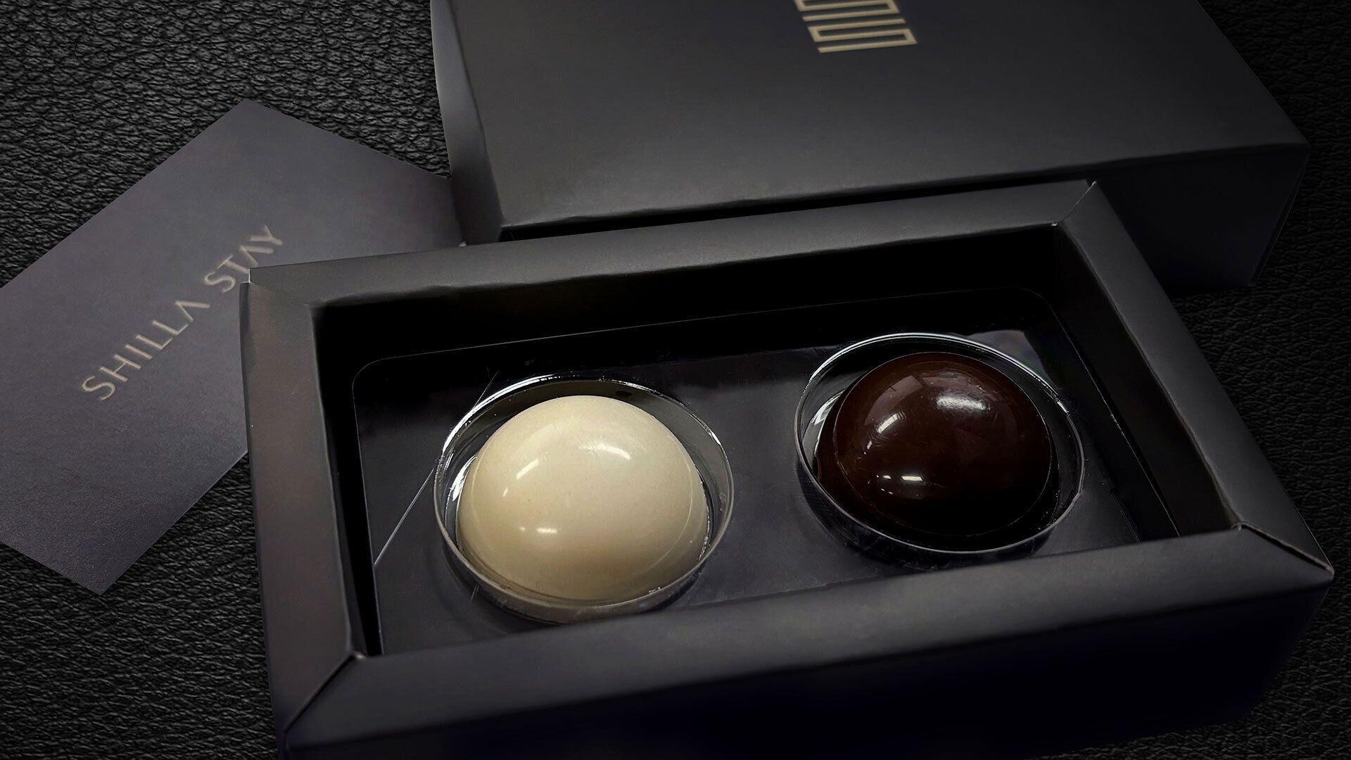 A black box containing two spherical objects, one white and one dark brown, with a label reading "SHILLA STY" on a textured surface.