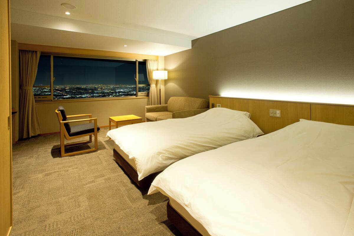Een hotelkamer met twee bedden, een stoel, een bureau en een raam met uitzicht op de stad.