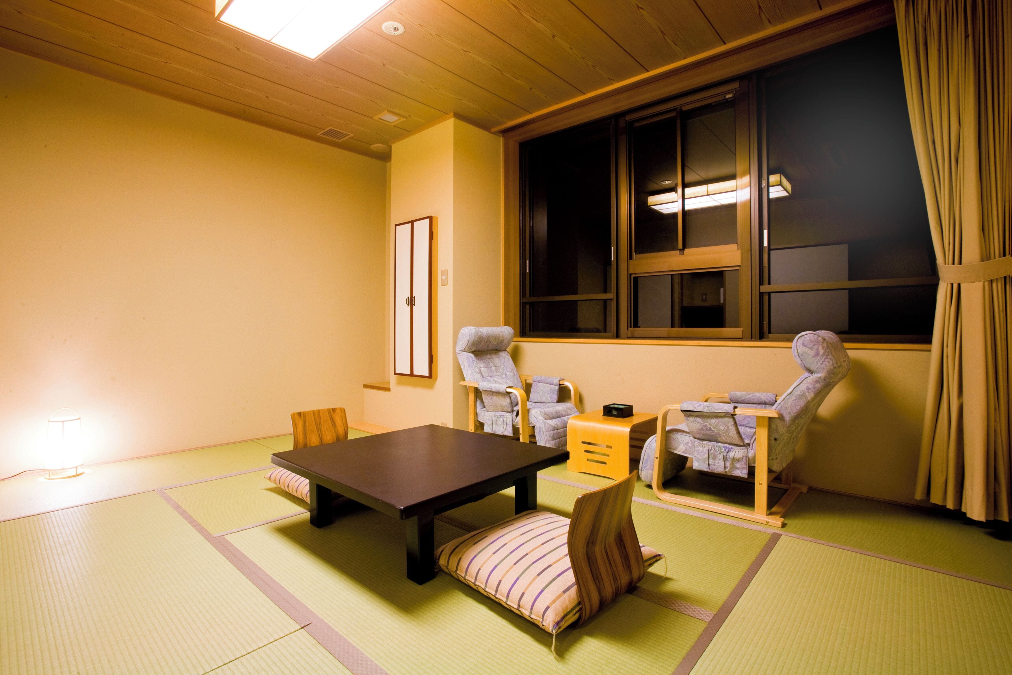 Une pièce de style japonais traditionnel, avec un sol recouvert de tatamis, une table basse, deux chaises, une petite table et une fenêtre avec des rideaux.