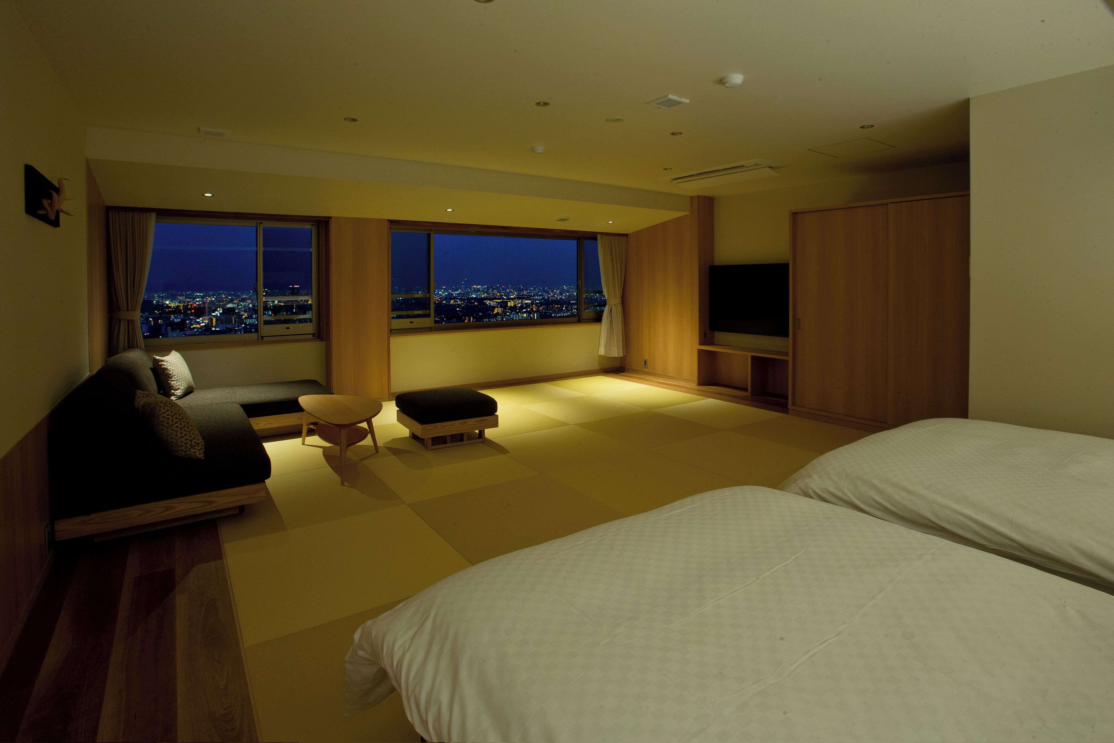 A modern hotel room with a large window offering a city view, a sofa, a small table, and a bed.