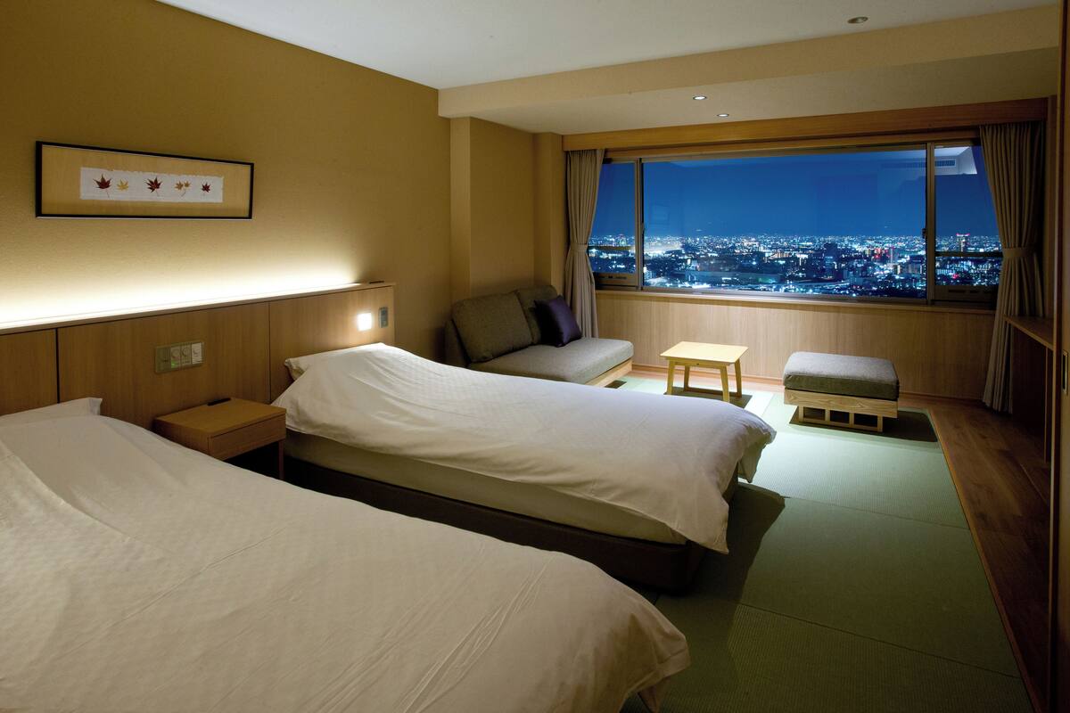 Hotelkamer met twee bedden, een bank, een tafeltje en uitzicht op een stadsbeeld in de nacht.