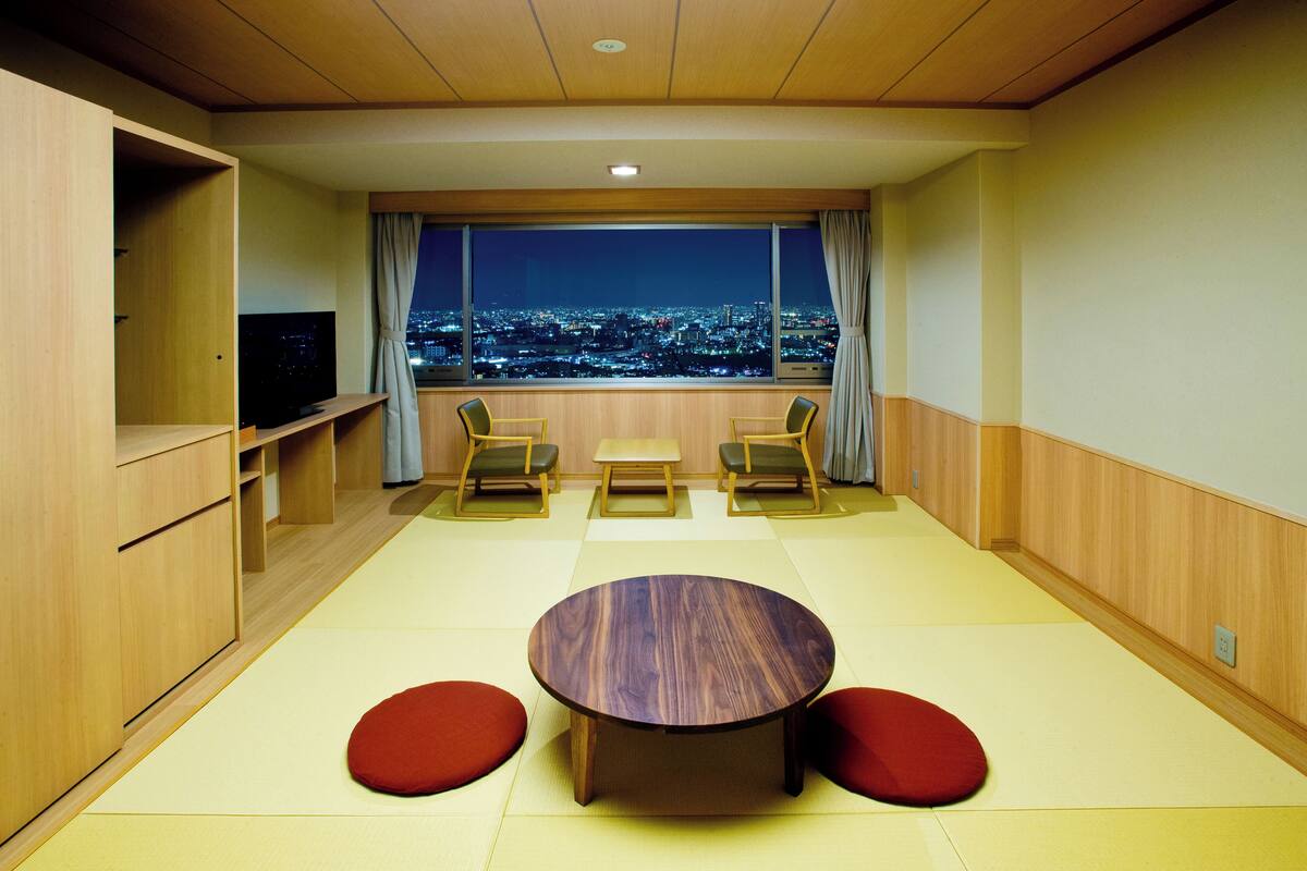 Een kamer met tatami vloer, een ronde tafel, twee stoelen, een televisie en uitzicht op een stadsbeeld 's nachts.