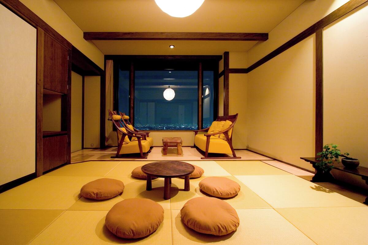 Een traditionele Japanse kamer met tatami vloeren, twee houten stoelen, een kleine tafel en uitzicht op de stad 's nachts.