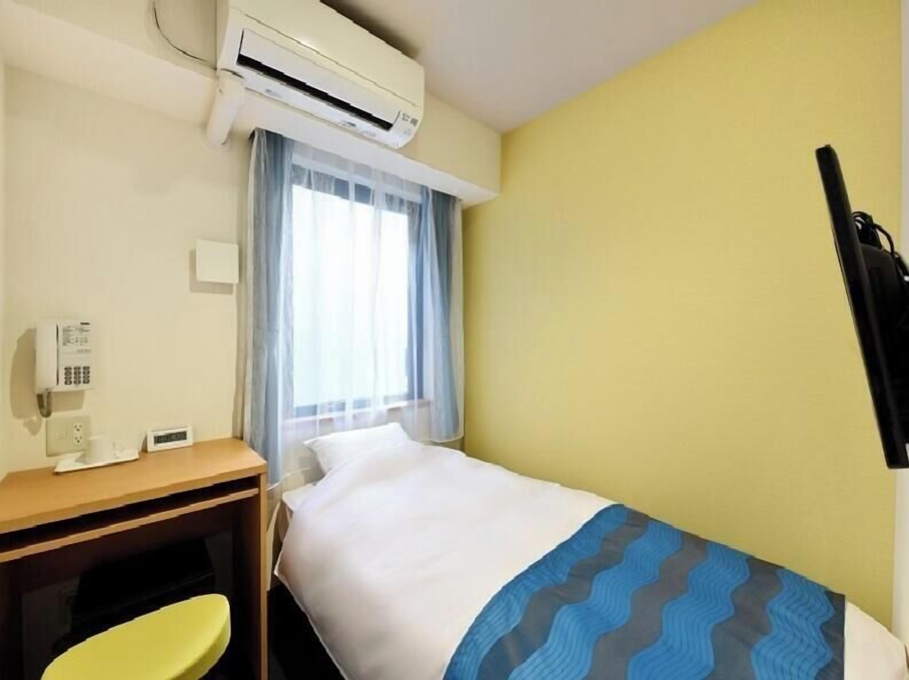 Hotellihuone, jossa on sänky, työpöytä, tuoli, ilmastointilaite ja ikkuna, jossa on verhot.