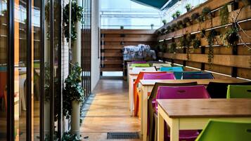 Un couloir avec des chaises et des tables colorées, des murs en bois ornés de plantes et une porte en verre sur la gauche.