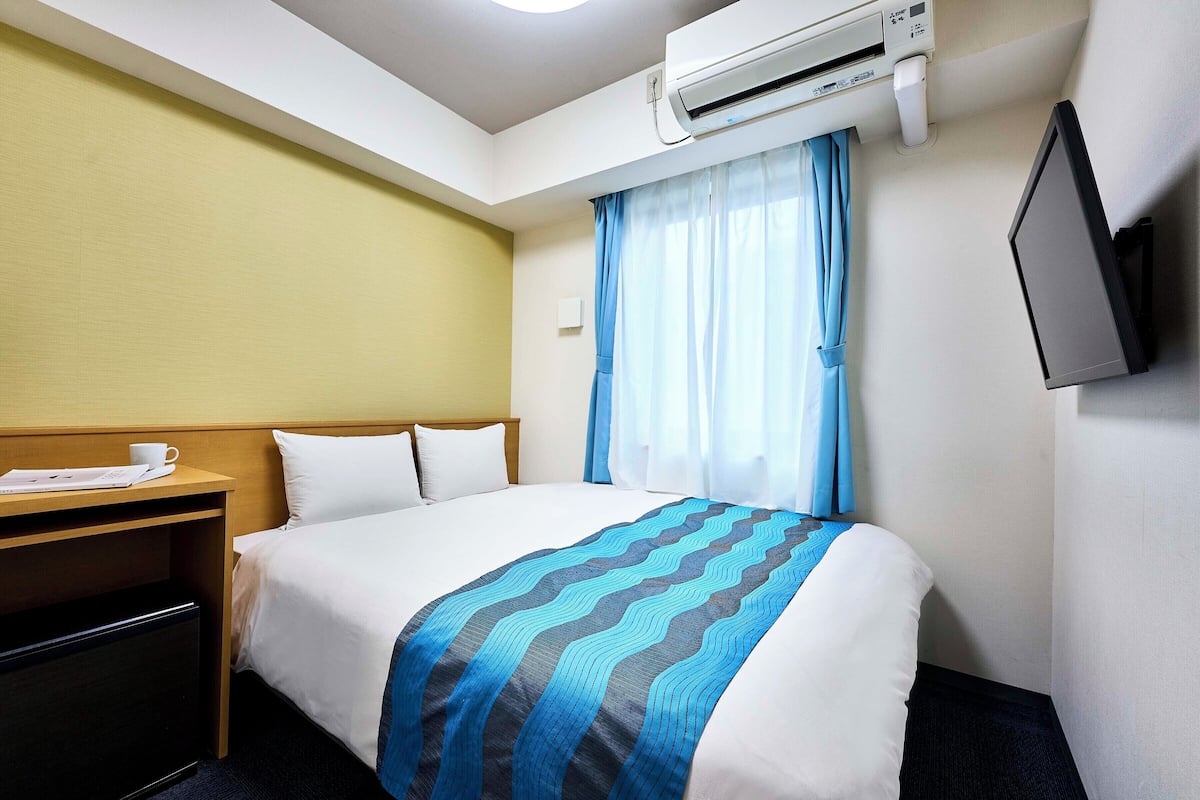 A hotel room with a bed, a desk with a cup and papers, an air conditioner, a TV, and a window with curtains.
