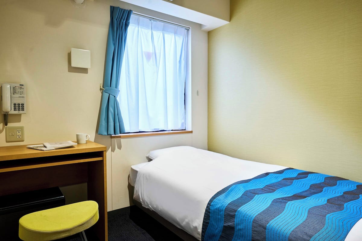 A small hotel room with a single bed, a desk with a telephone, a chair, and a window with curtains.