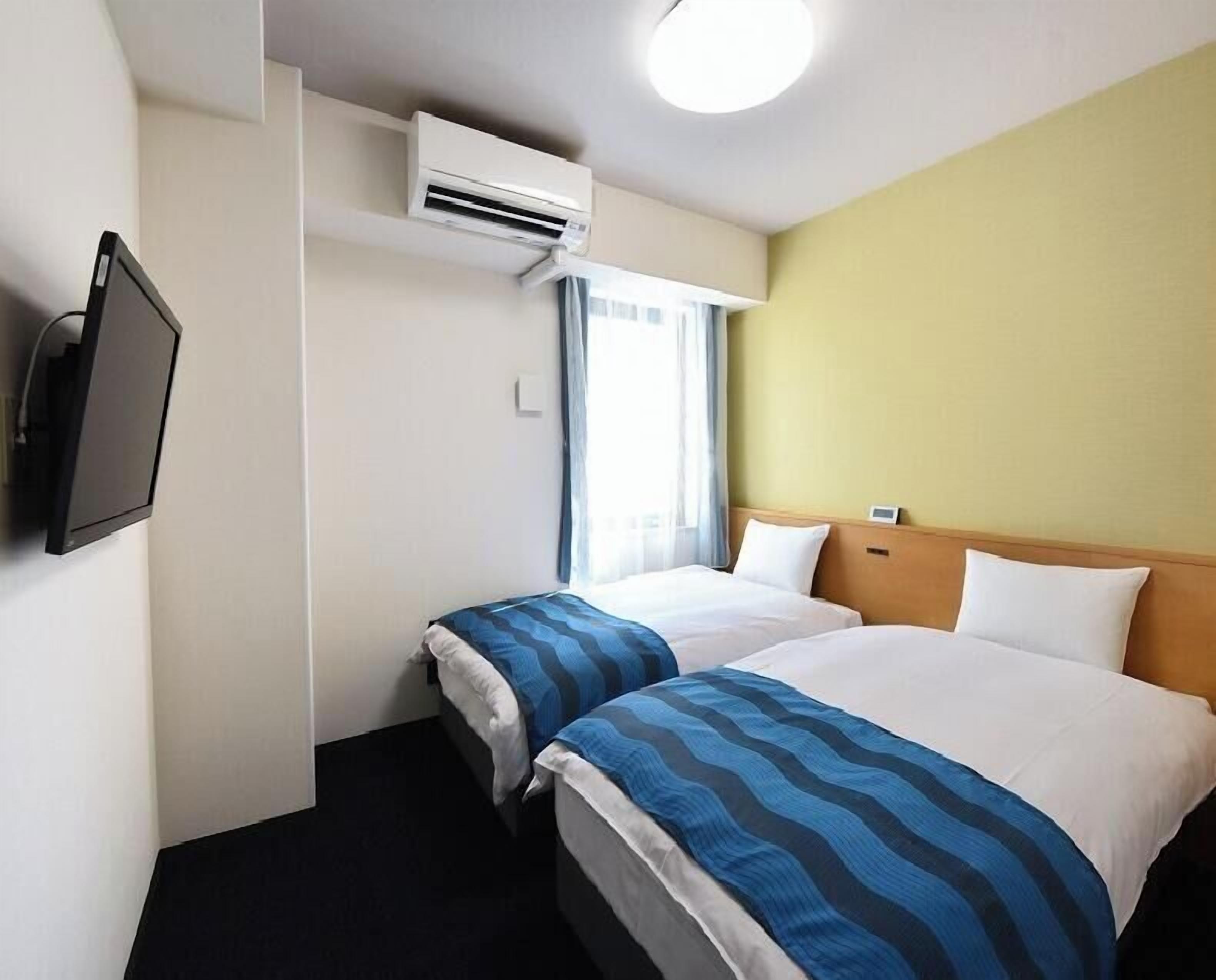 Hotellihuone, jossa on kaksi sänkyä, ilmastointilaite, televisio ja ikkuna, jossa on verhot.