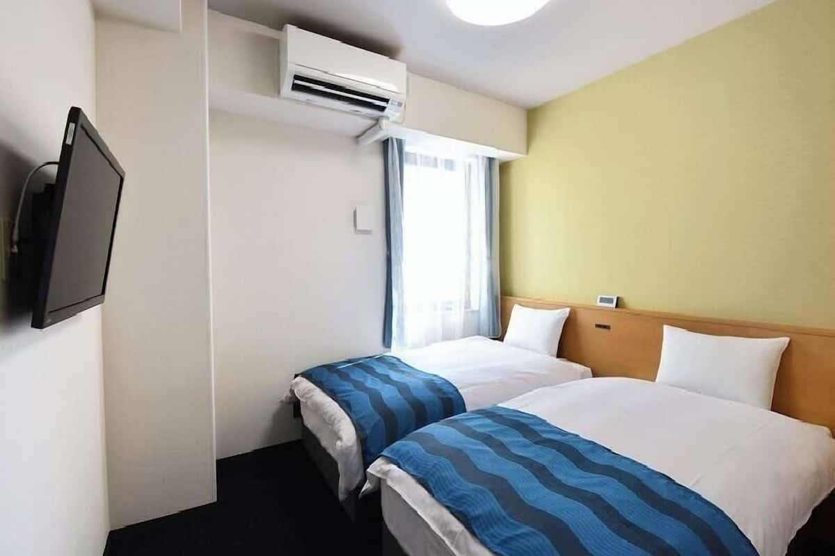 A hotel room with two beds, an air conditioning unit, a television, and a window with curtains.