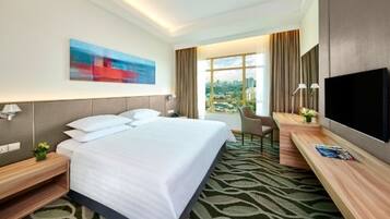 A hotel room with a large bed, a desk with a TV, a chair, and a window with a city view.