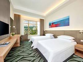 Habitación de hotel con dos camas, un escritorio, un televisor y un ventanal con vistas a la ciudad.