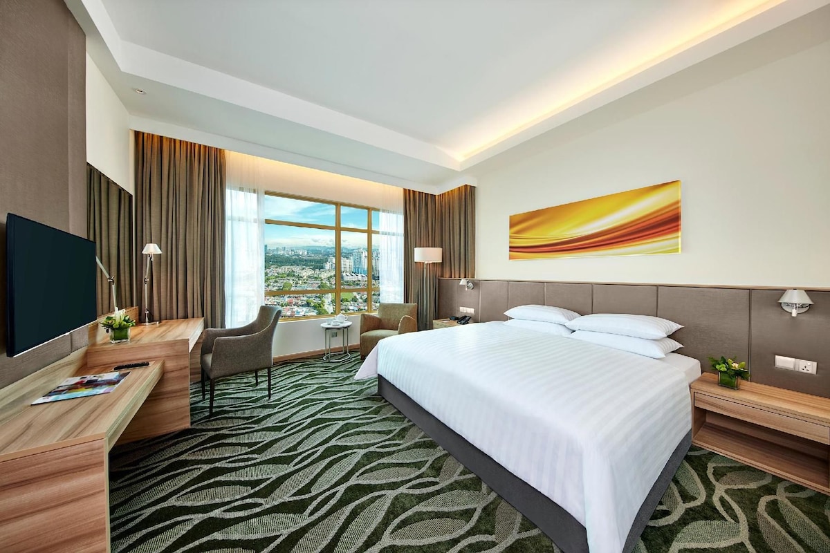 Een hotelkamer met een bed, bureau, stoel en een groot raam met uitzicht op de stad.