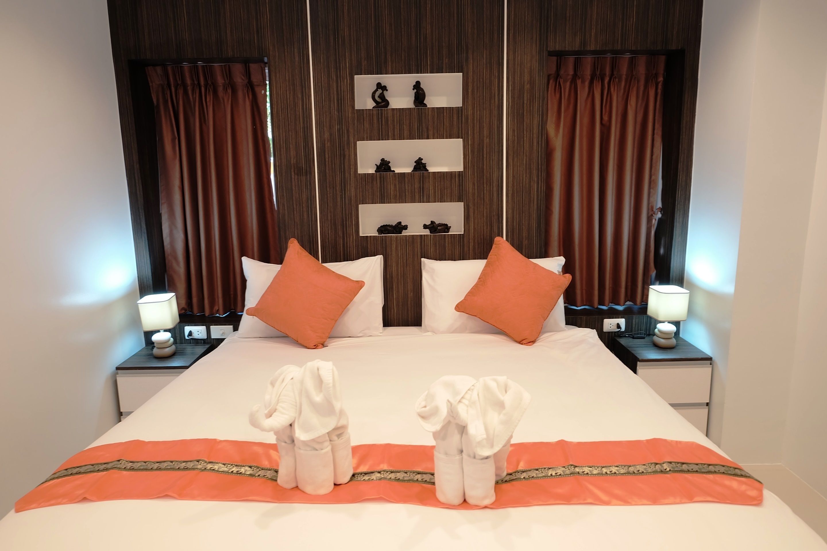 A neatly made bed with white and orange bedding, two nightstands with lamps, and a wall with three shelves displaying figurines.