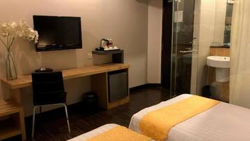 A hotel room with two beds, a desk with a TV, a small fridge, a chair, and a shower area.