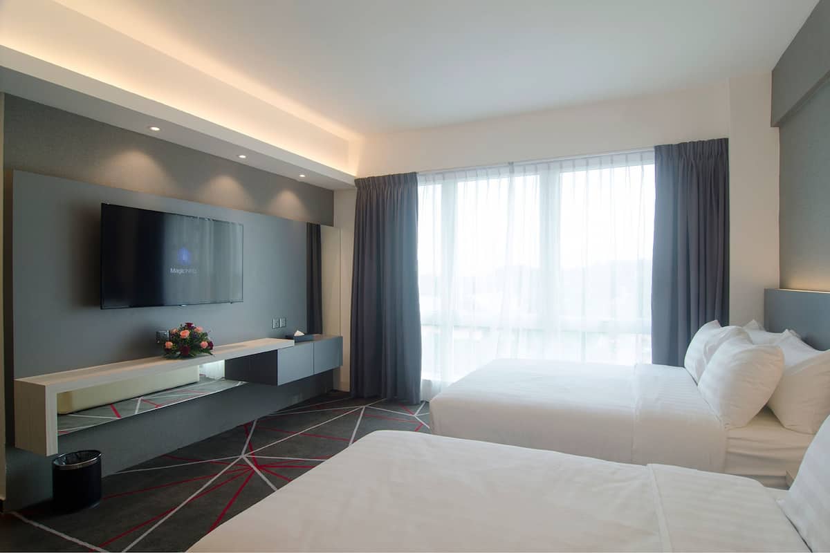 Habitación de hotel con dos camas, un televisor en la pared, una mesita con un jarrón de flores y un ventanal grande con cortinas.