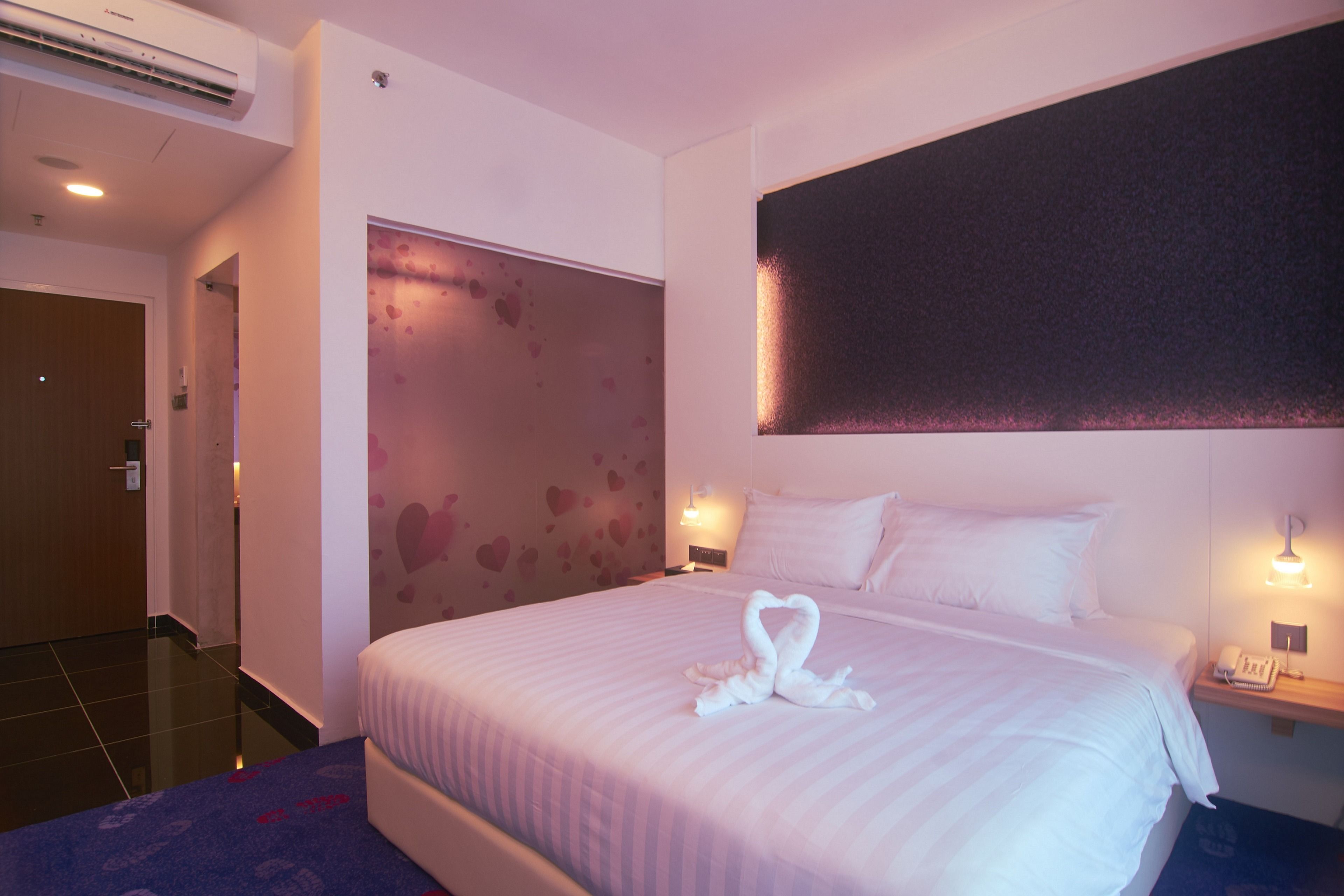 A hotel room with a large bed, a wall-mounted air conditioner, a bedside table with a lamp, and a door leading to another room.