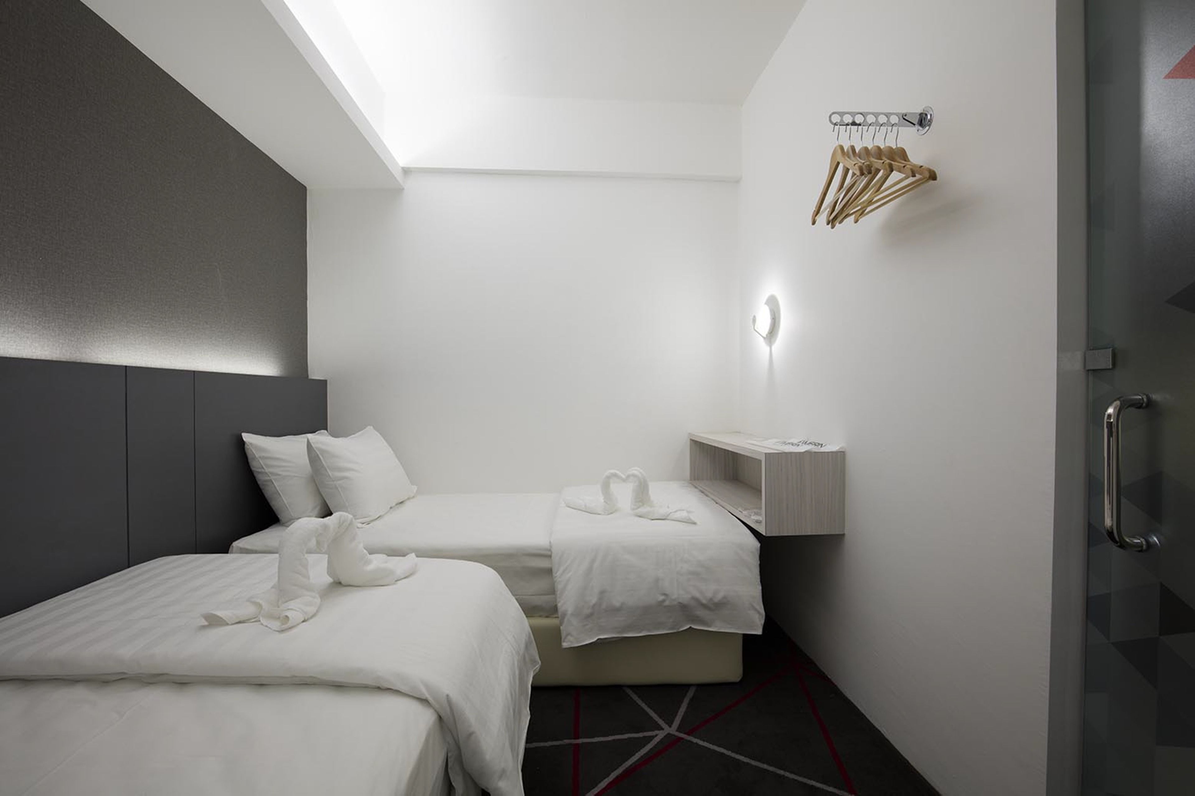 A modern hotel room with two beds, a small bedside table, a wall-mounted towel rack, and a glass door.
