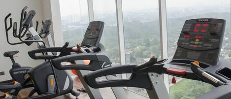 A gym with multiple exercise machines, including treadmills and stationary bikes, with a large window offering a city view.