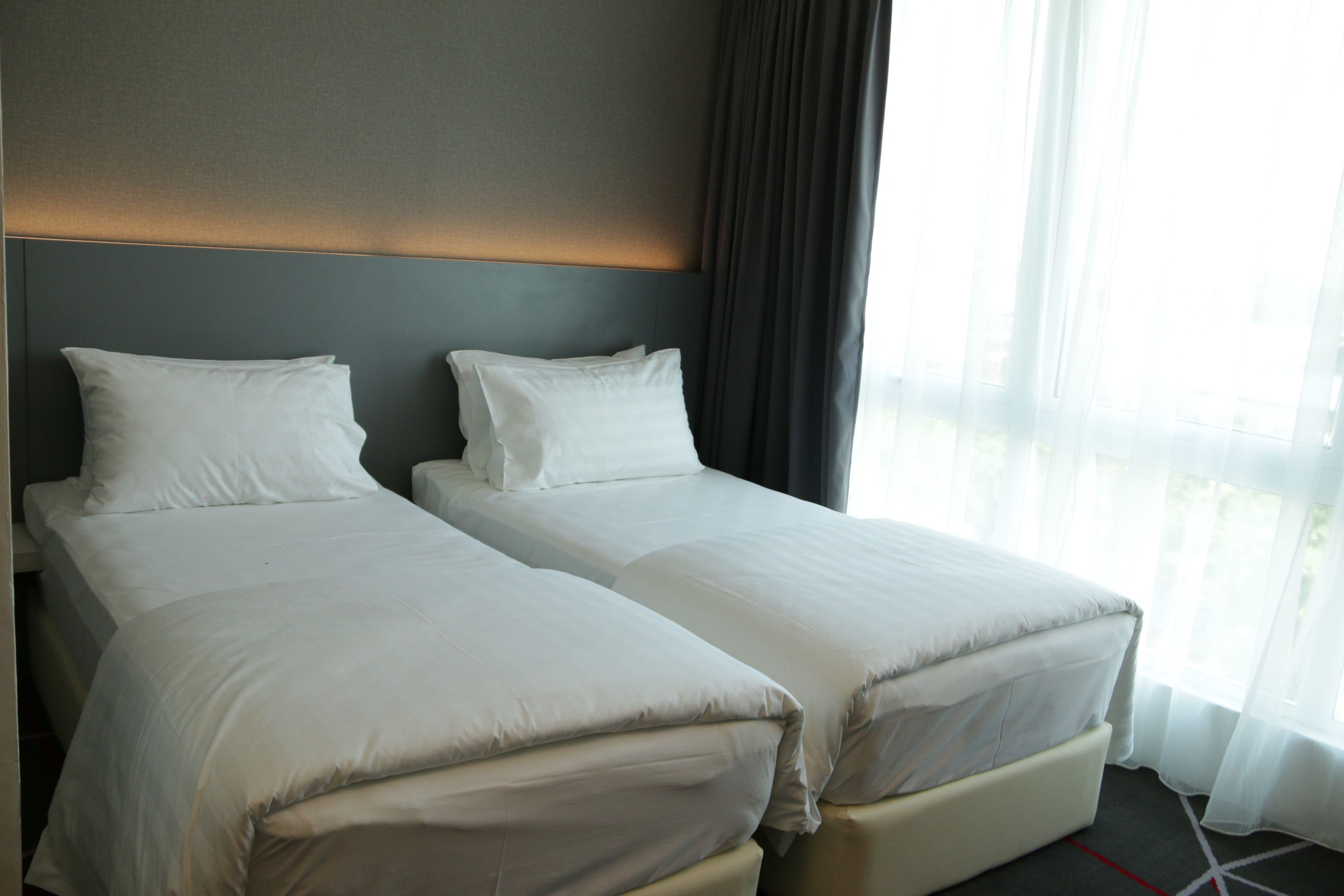 A hotel room with two single beds, a headboard with integrated lighting, and a large window with sheer curtains.