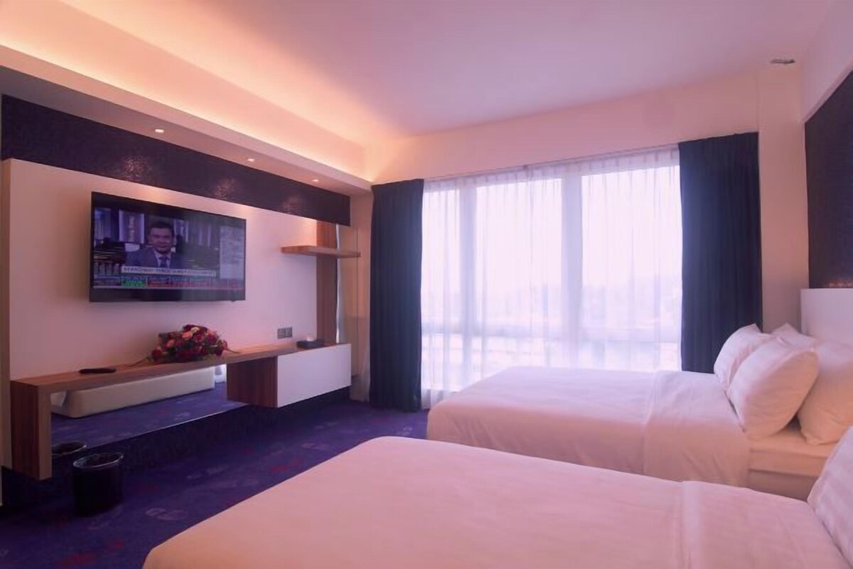 Habitación de hotel con dos camas, un televisor de pantalla plana montado en la pared, una mesita con un florero y un ventanal con cortinas ligeras.