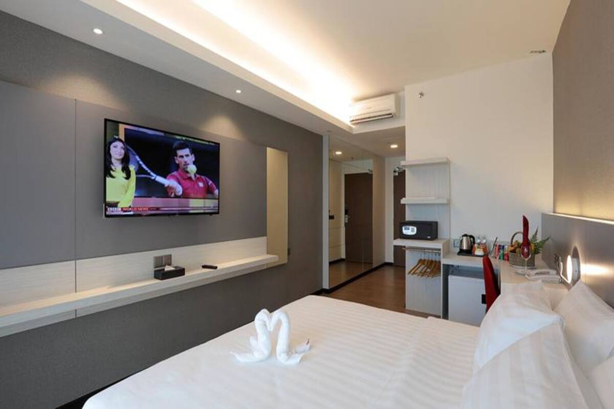 Habitación de hotel moderna con una cama grande, un televisor de pantalla plana en la pared, un escritorio pequeño con una lámpara y un estante en la pared.