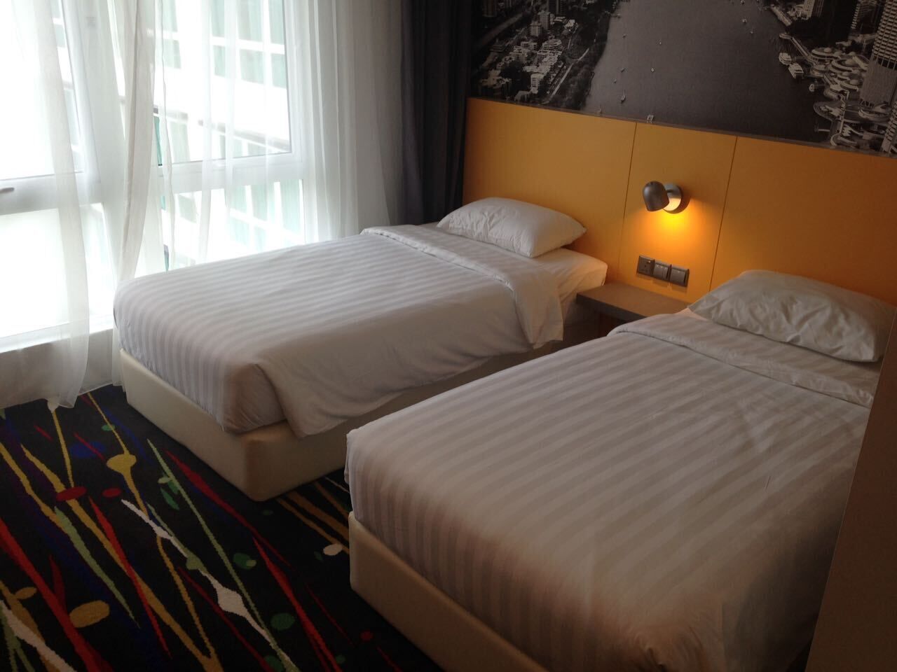 Una habitación de hotel con dos camas individuales, una mesita de noche con lámpara y una alfombra abstracta de colores.