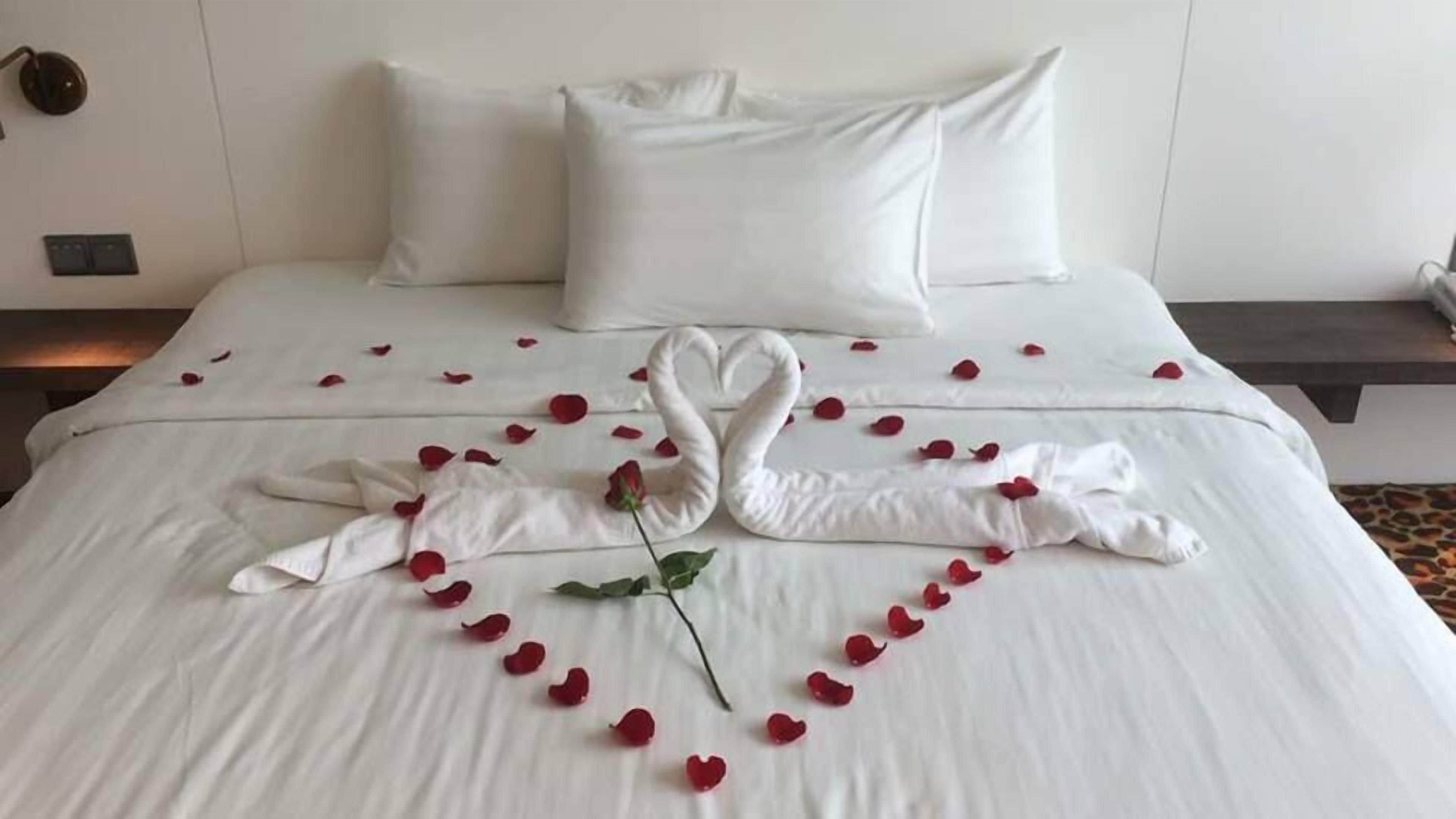 A neatly made bed with white linens, a heart-shaped arrangement of rose petals, and two swan-shaped pillows.