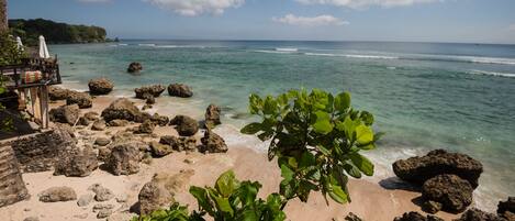 A beach with rocky terrain, clear turquoise water, and a wooden structure with umbrellas.