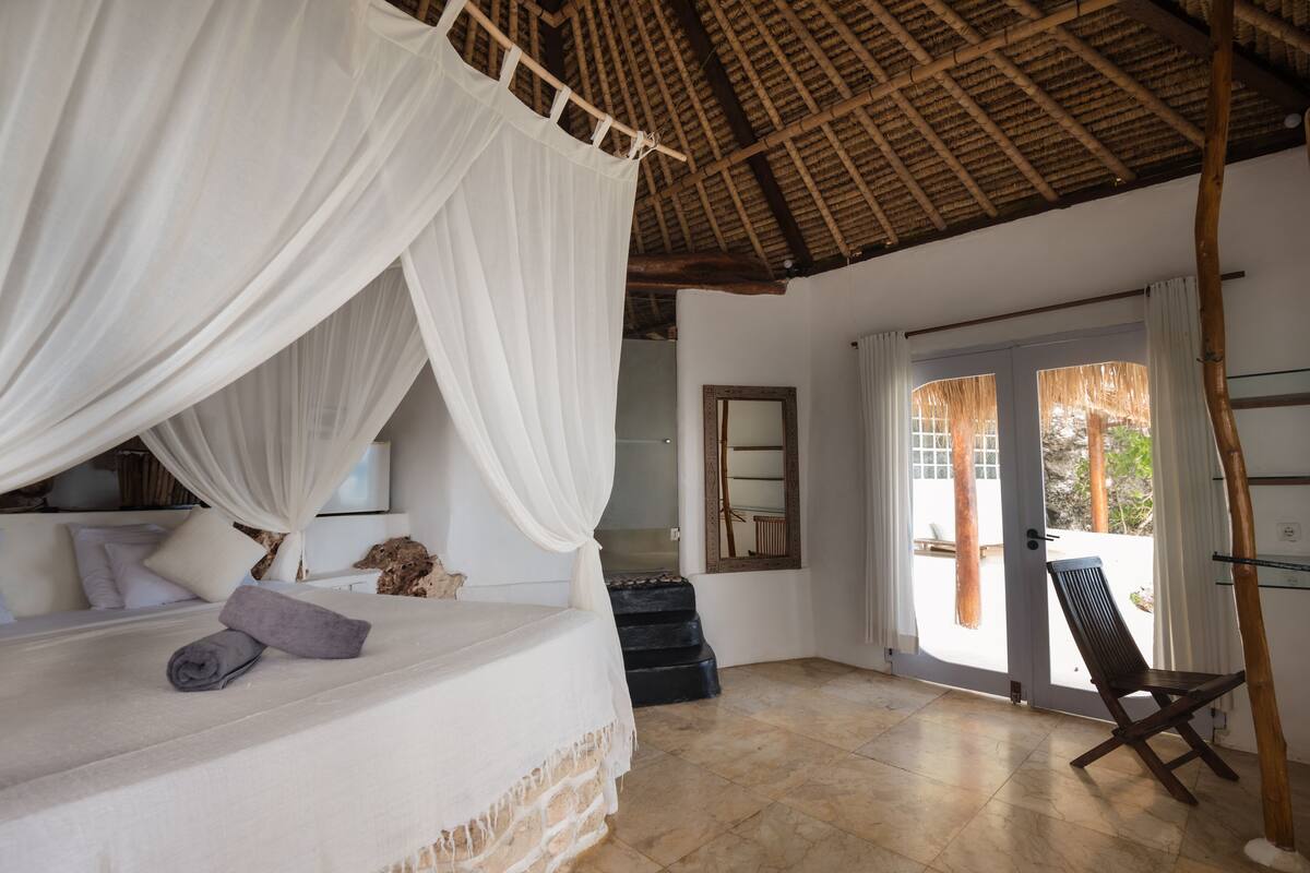 A bedroom with a canopy bed, a wooden ceiling, a view through a glass door, and a chair by the door.