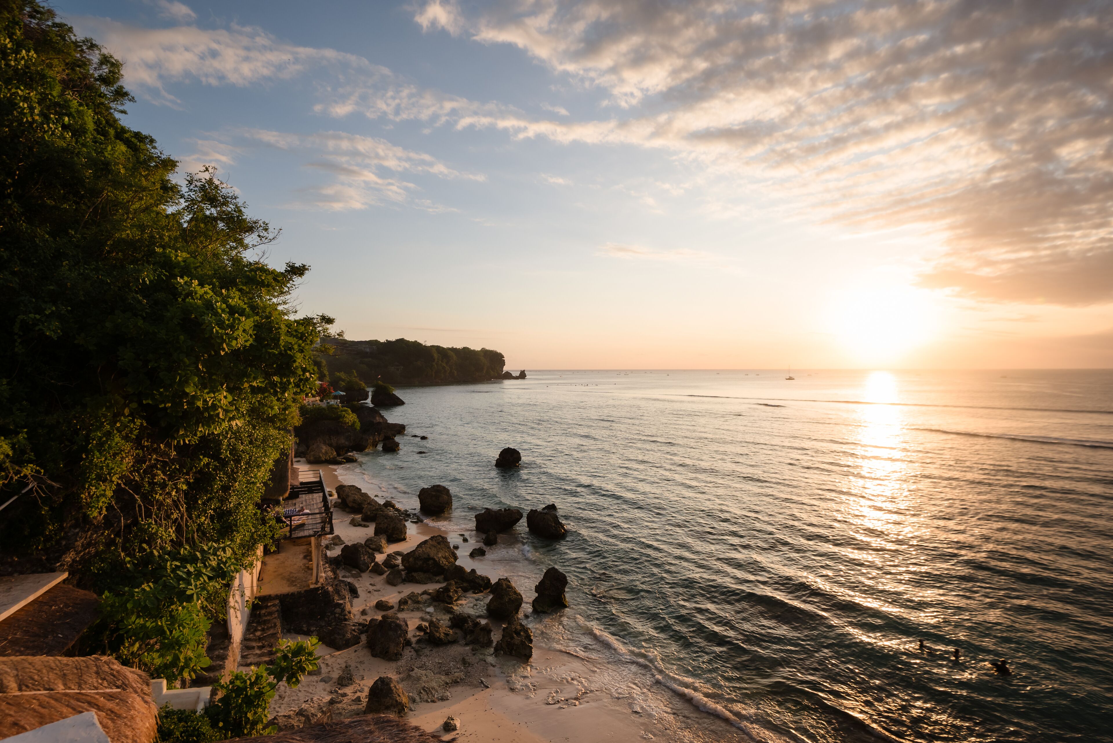 A coastal view with a rocky shoreline, a clear sky, and the sun setting over the water.