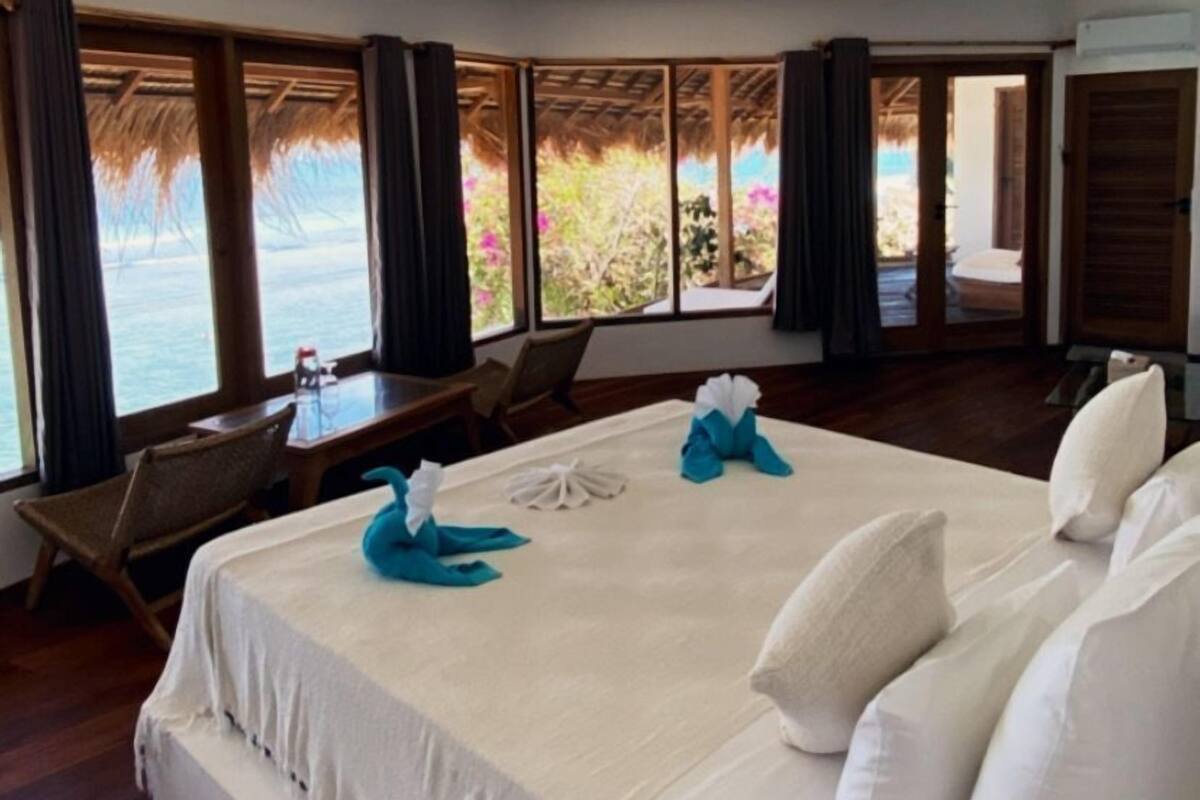 A spacious bedroom with a large bed, a wooden floor, and a view of the sea through large windows.