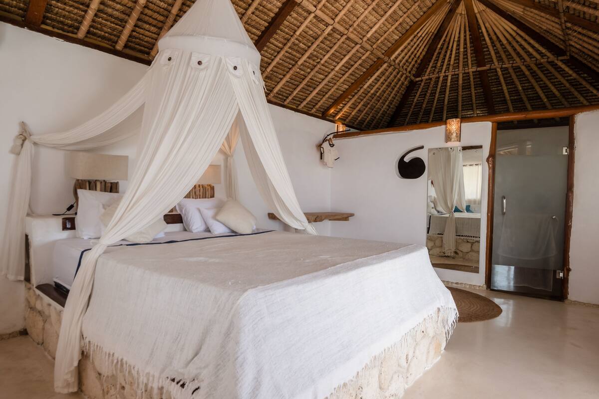 A spacious bedroom with a large bed, a canopy, a stone wall, and a thatched roof.