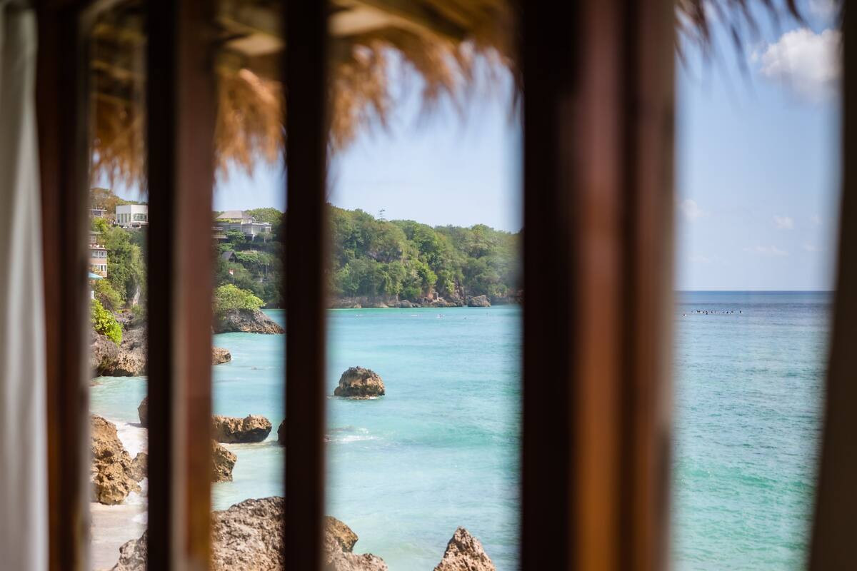 A coastal view through a window with a thatched roof, rocky shoreline, and clear turquoise water.