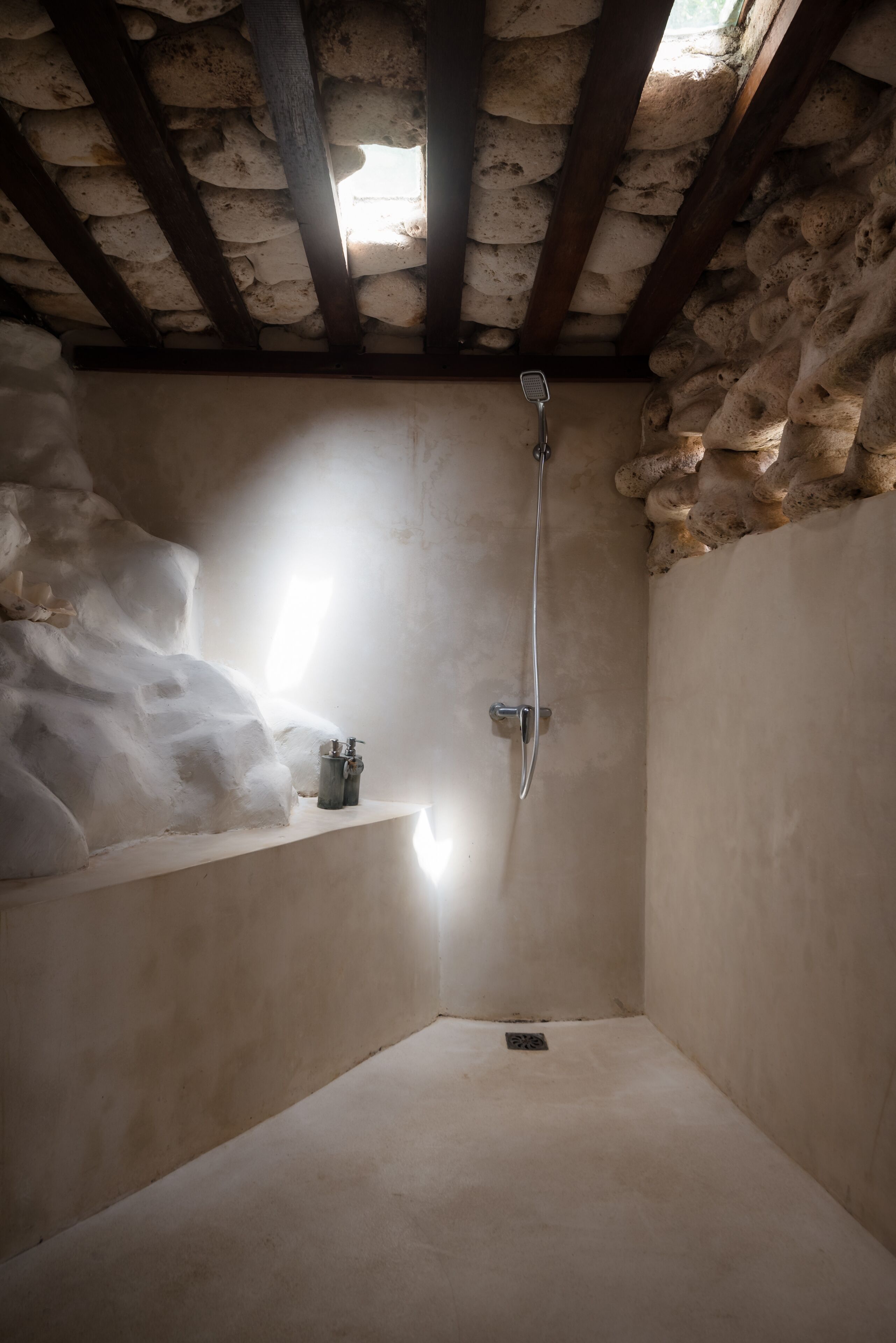 En bruseniche med stenvægge, loftsbjælker af træ, et håndholdt brusehoved og en lille hylde med toiletartikler.