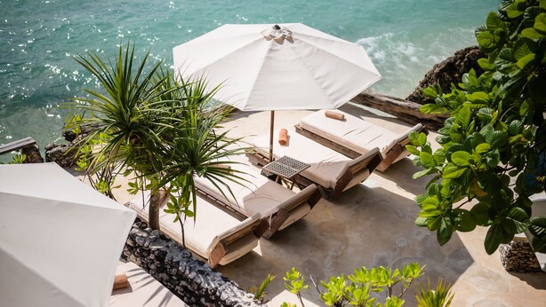 A seaside lounge area with sun loungers, a white umbrella, and a palm tree. - Rock 'n Reef Hotel (Pecatu)