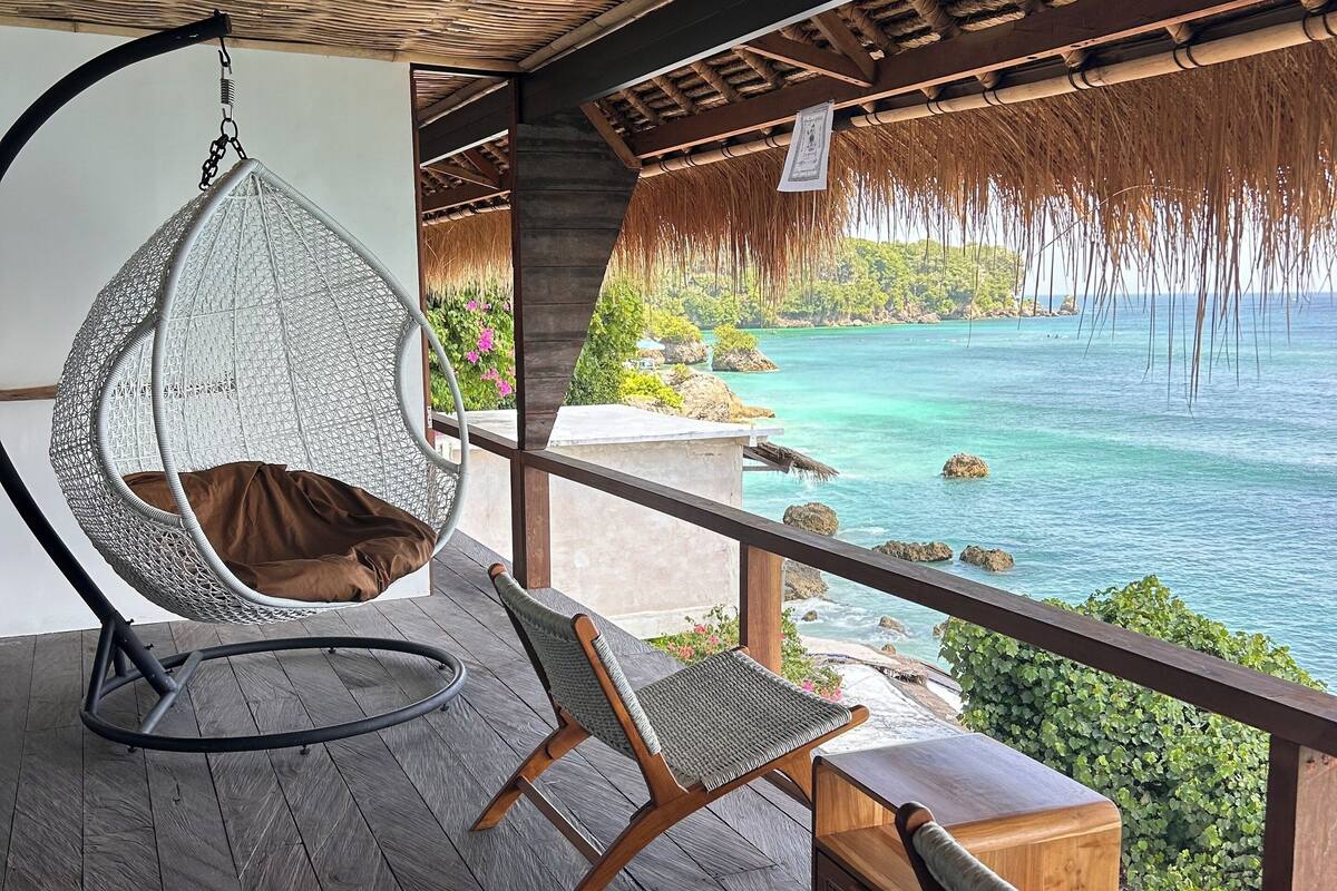 A wooden deck with a hanging chair and two wooden chairs, overlooking a clear blue sea and green landscape.