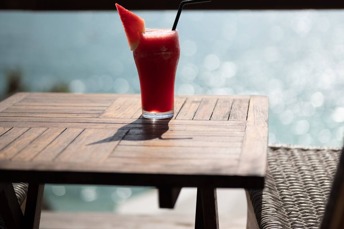 A wooden table with a glass of red drink, a straw, and a slice of fruit on a balcony overlooking a body of water.