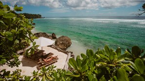 A beach view with a lounge area, clear turquoise water, and rocky outcrops. - Rock 'n Reef Hotel (Pecatu)