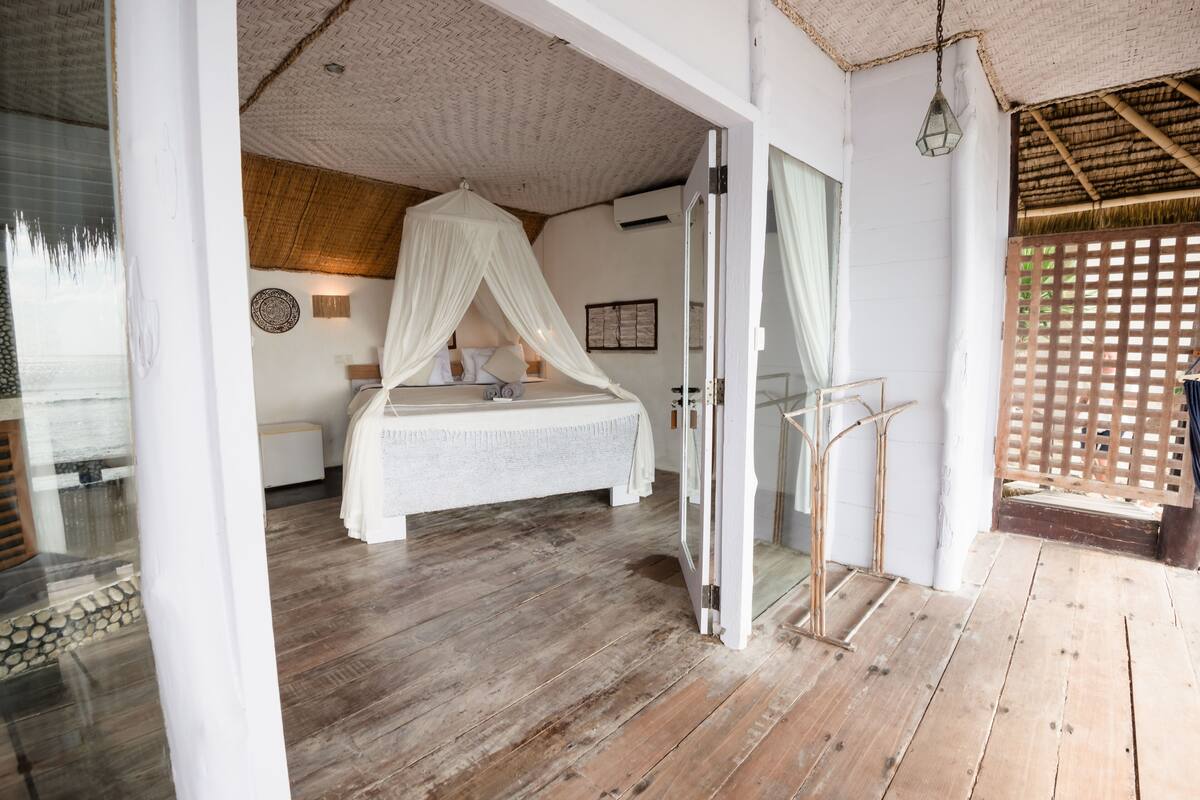 A bedroom with a bed under a canopy, wooden flooring, a glass door leading outside, and a view of a wooden fence.