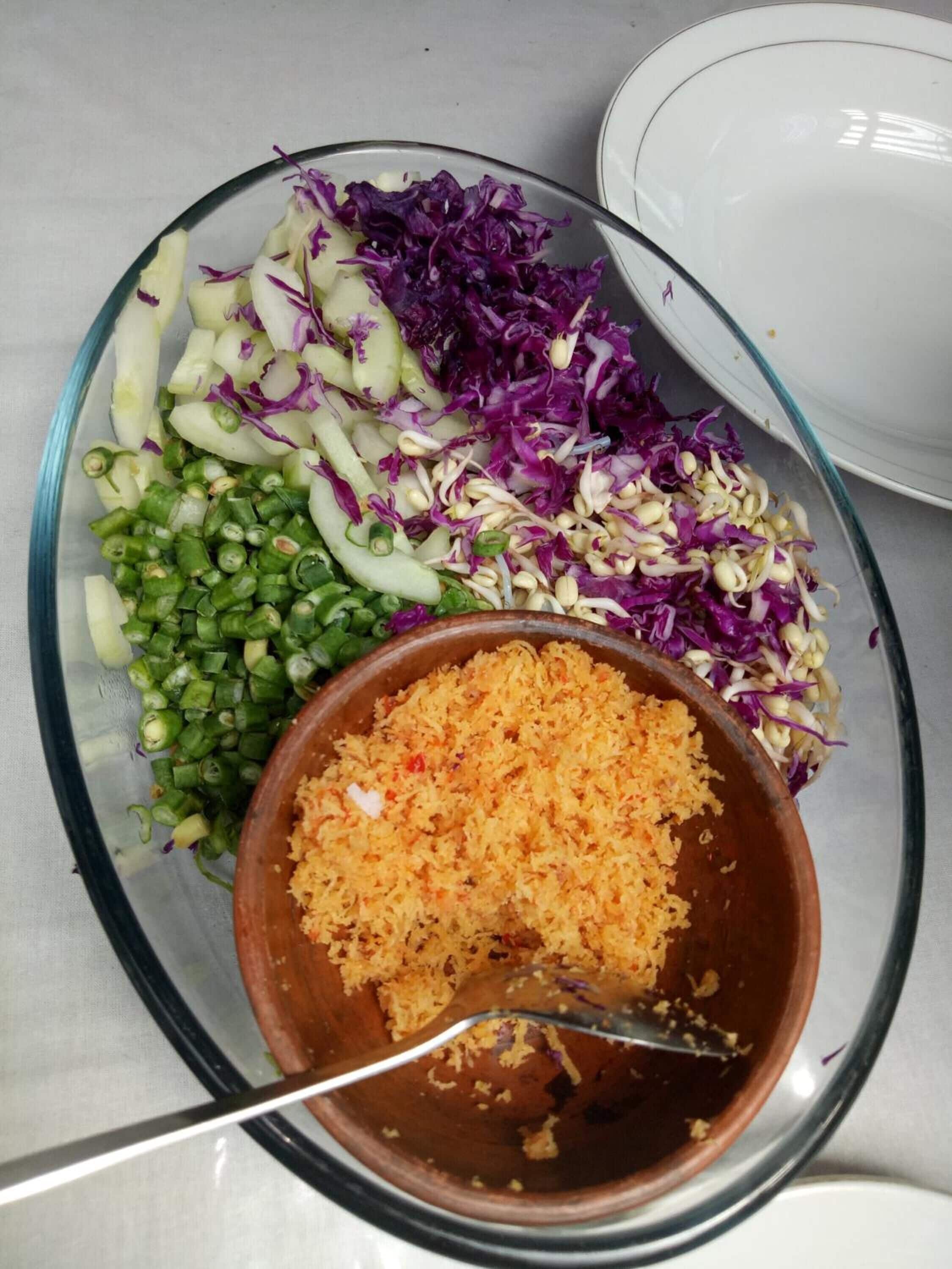 Un cuenco de vidrio con una ensalada de diversos vegetales y un recipiente pequeño con una mezcla amarillenta, posiblemente aderezo o condimento, sobre una mesa.