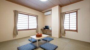 A room with tatami mat flooring, a low table with cushions, a small desk with a computer, and two windows with curtains.