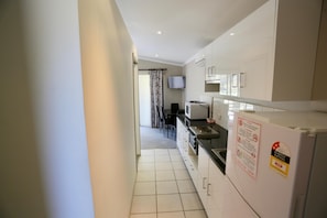 A narrow kitchen with white cabinets, a refrigerator, and a television mounted on the wall. - Junee Tourist Park (Junee)