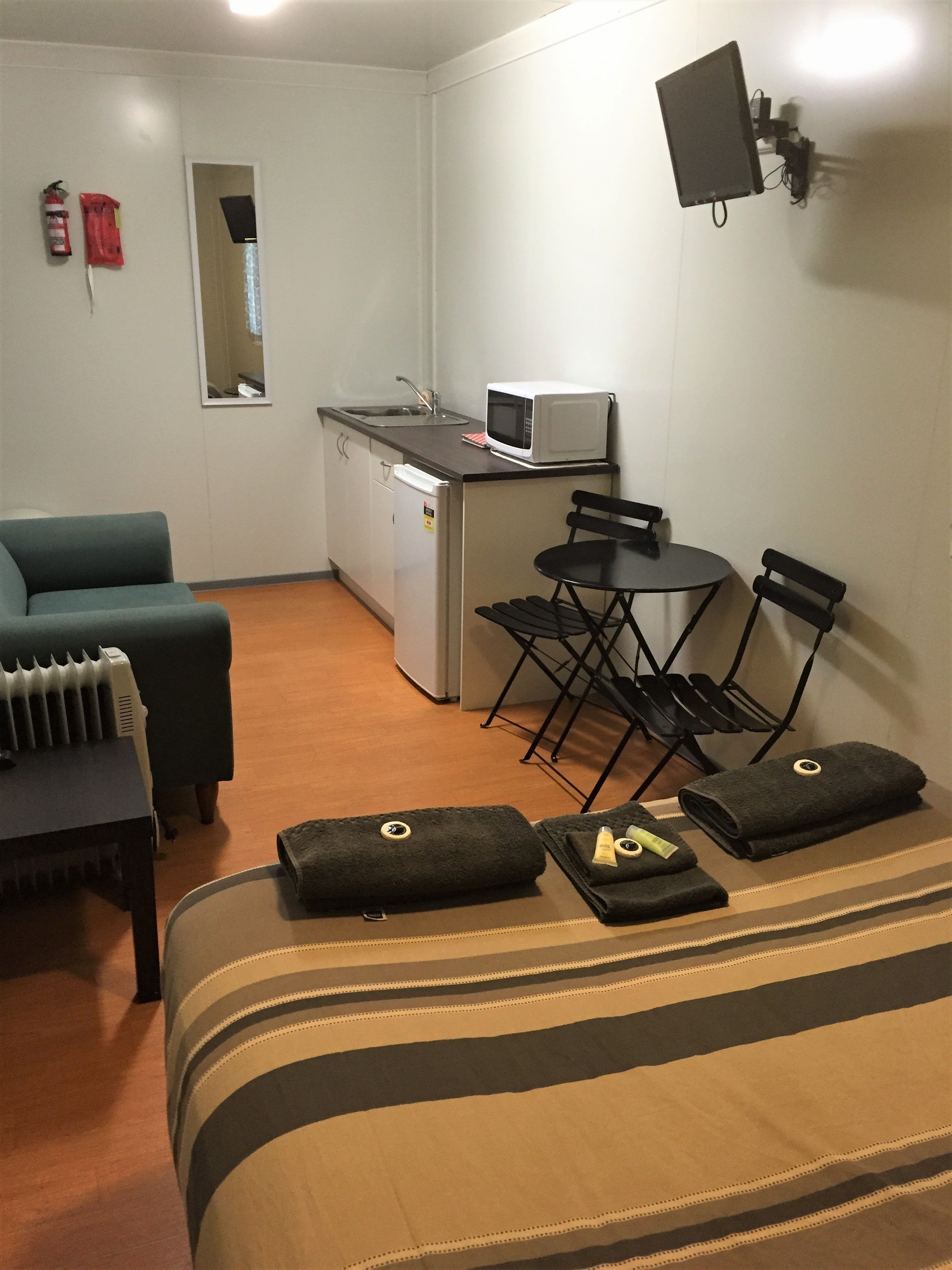 A compact hotel room with a kitchenette, a sofa, a small dining table with chairs, and a flat-screen TV mounted on the wall.