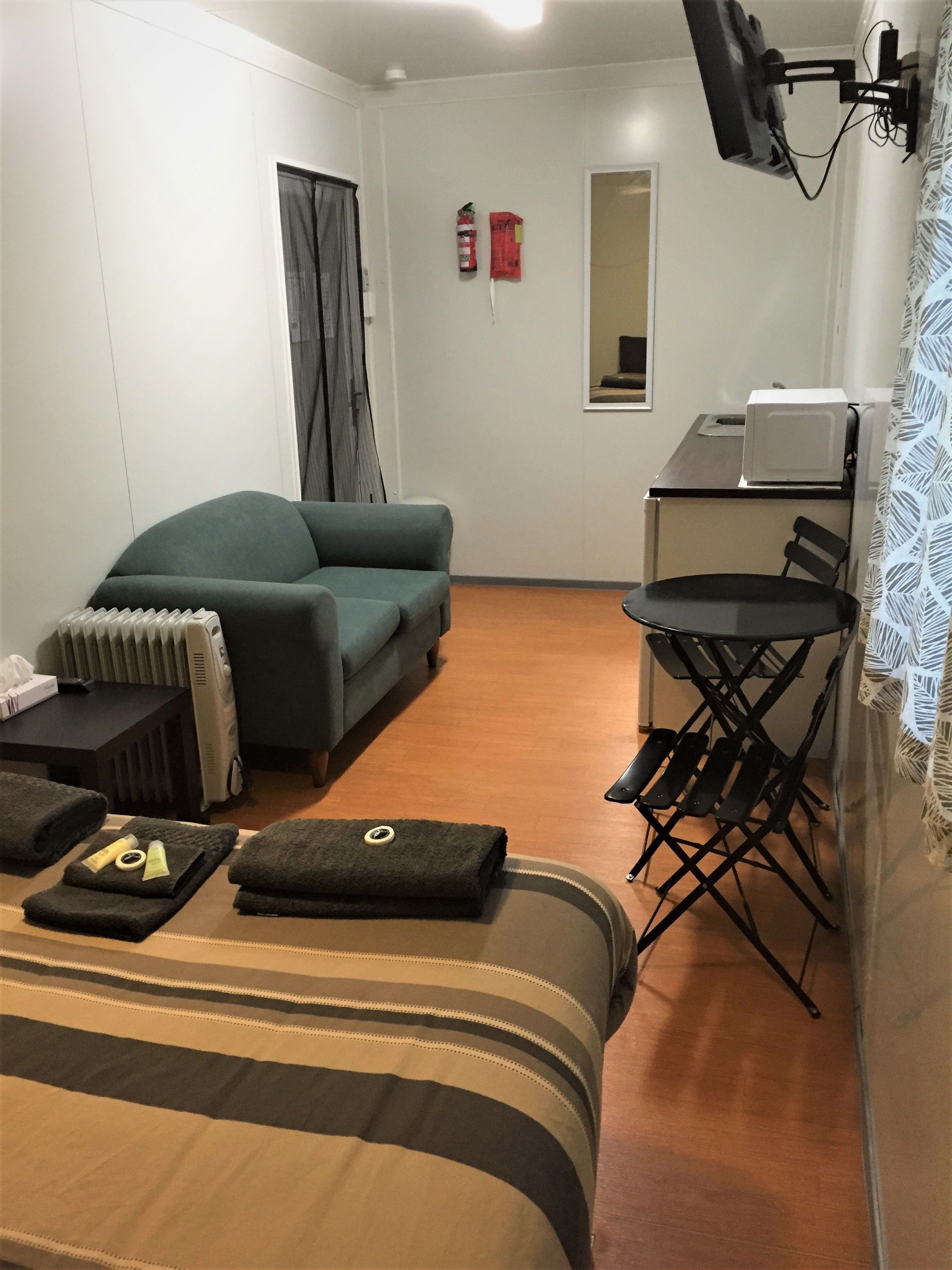 A hotel room with a sofa, a small table with a radiator, a foldable chair, a microwave, and a flat-screen TV mounted on the wall.