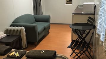 A hotel room with a sofa, a small table with a radiator, a foldable chair, a microwave, and a flat-screen TV mounted on the wall.