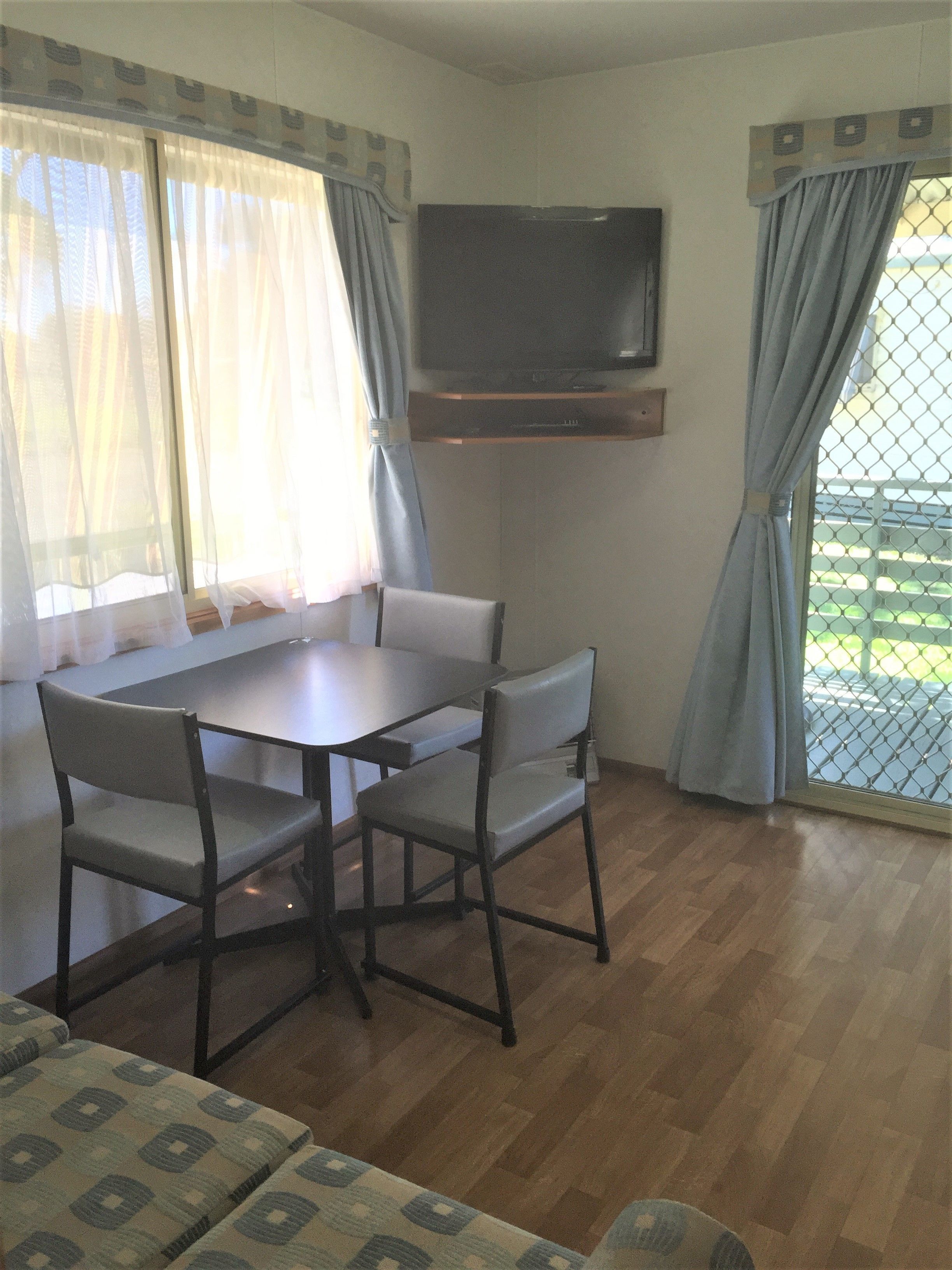 A room with a dining table, four chairs, a television mounted on the wall, and a window with curtains.