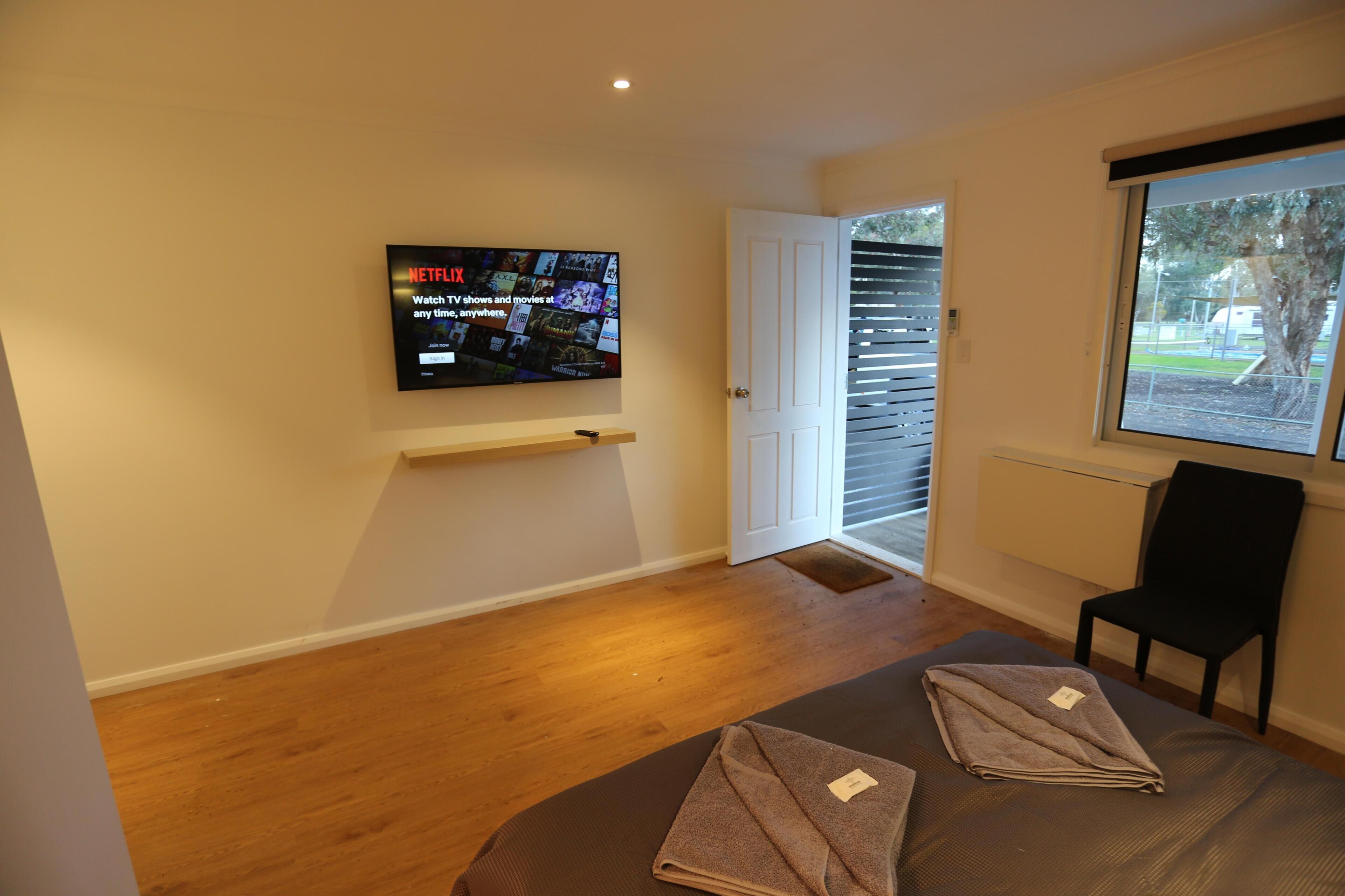 A modern room with a flat-screen TV mounted on the wall, a door leading to an outdoor area, a window with blinds, and a chair.
