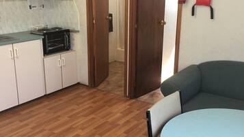 Cocina con armarios blancos, microondas y estufa. El suelo es de madera y hay una puerta que da al baño.
