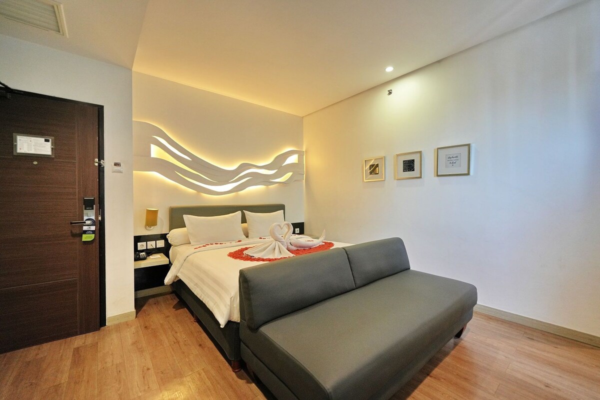 Une chambre d’hôtel moderne avec un lit, un canapé gris, un plancher en bois et un mur orné de tableaux encadrés.